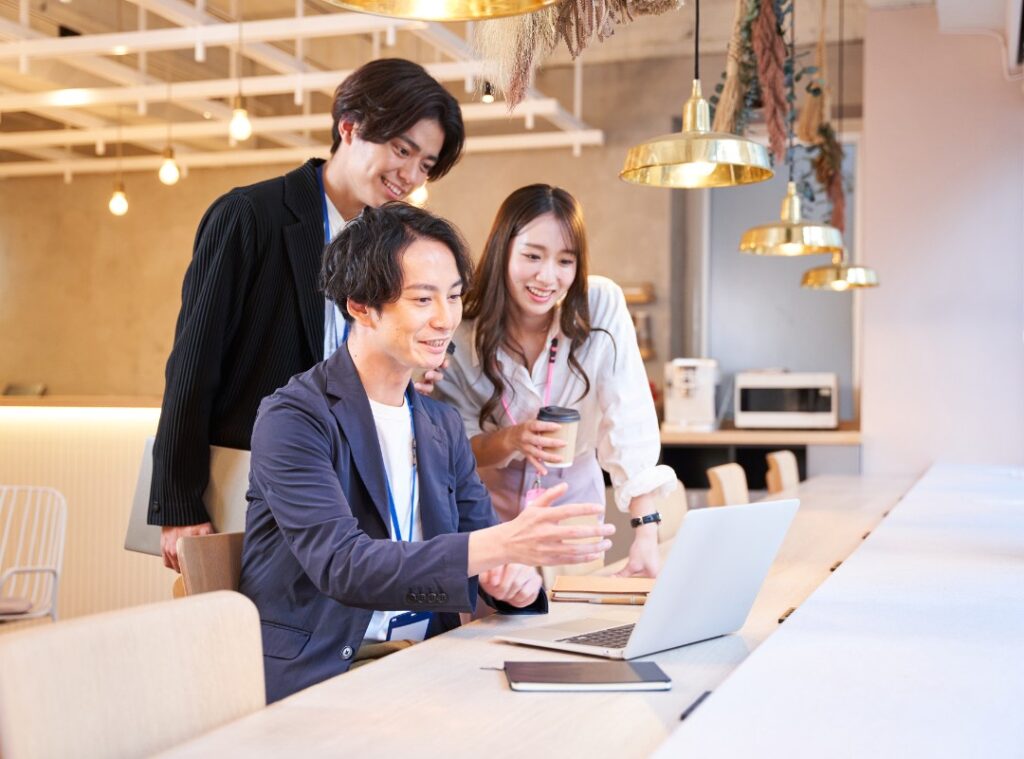 貸しオフィスの会議室にて、壁面の大型モニターに映るリモート参加者とハイブリッド会議を行う4人のビジネスパーソン。